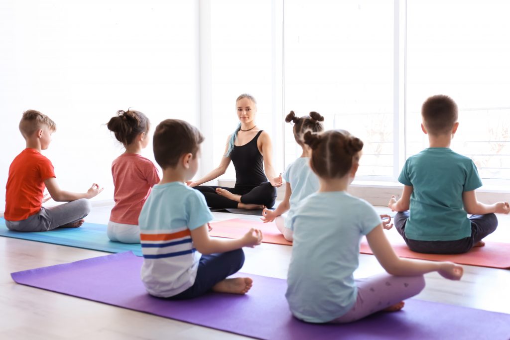 students sitting on mats facing yoga teacher, sitting in easy seated yoga pose with hands in a mindfulness mudra on knees