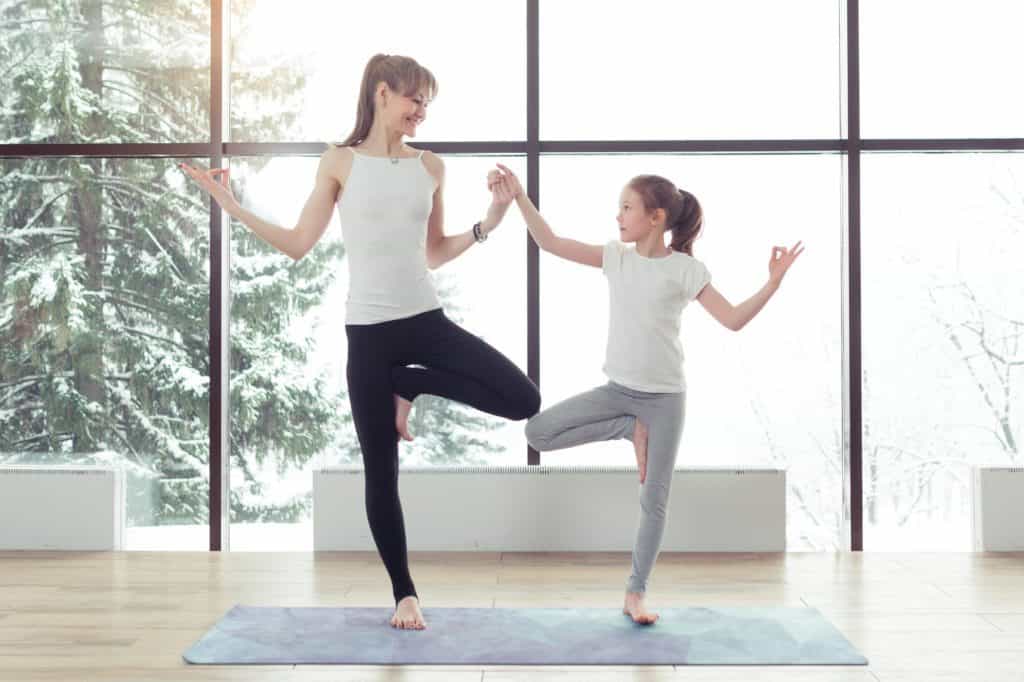 adulta and child are doing tree pose on a grew mat in front of a window, they are holding hands and looking at each other