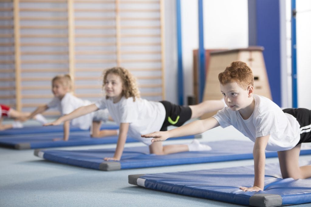 Three kids balance on one knee and opposite hand in a gymnasium on mats