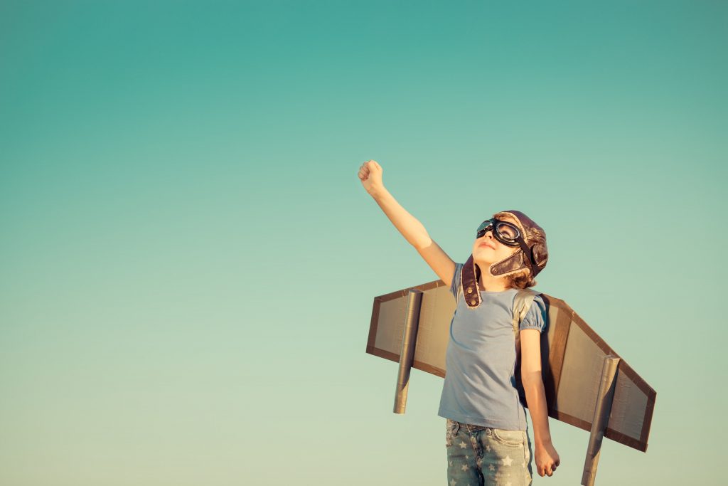 a young girl is dressed with a pilot mask and hat with a cardboard set of wings, she is lifting one arm and looking to the sky against a blue green sky background